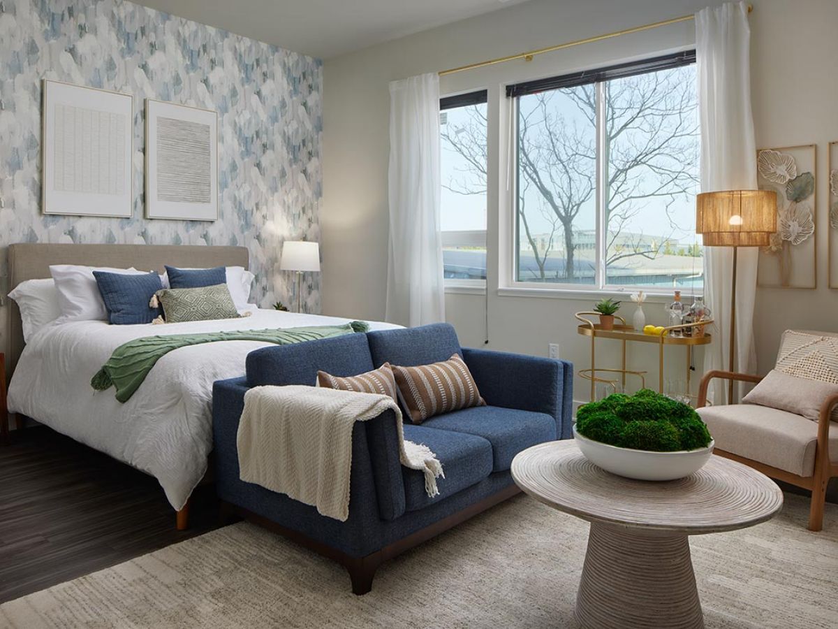 Westrum Corporate Modern bedroom at Luxor Wilmington with a blue loveseat, round table, large window, and accent wall—explore Wilmington apartments today.