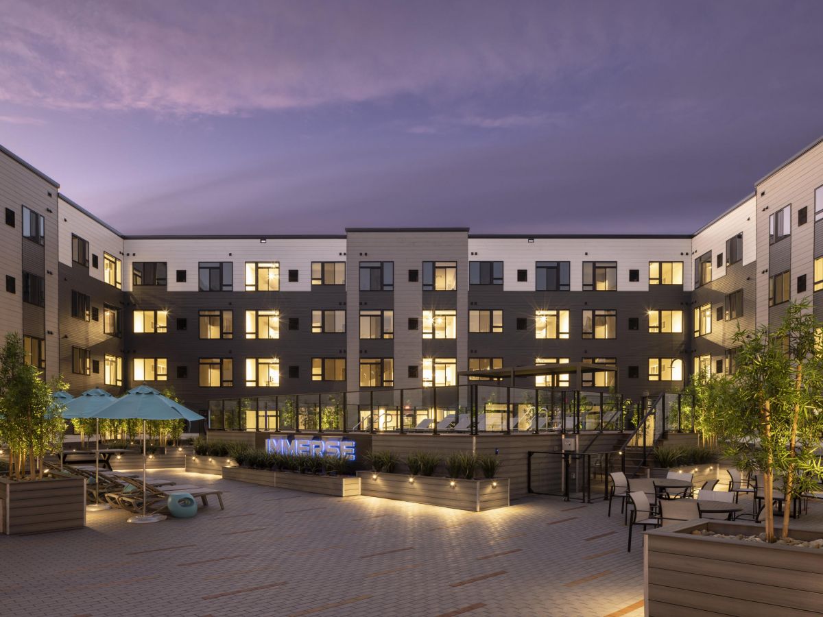 Westrum Corporate Modern apartment courtyard at dusk at Luxor Montgomeryville, featuring string lights, lush plants, and a glowing "IMMERSE" sign.