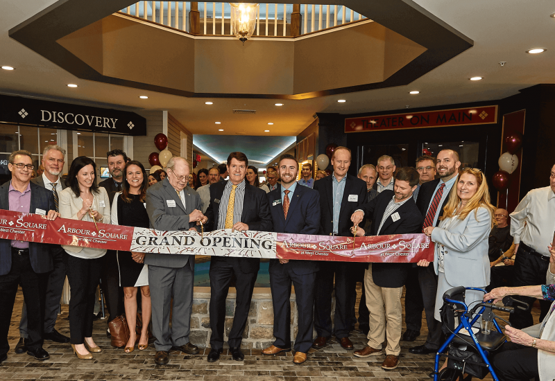 Westrum Corporate A group of people in business attire hold a ribbon that reads "Grand Opening" inside a building, with a crowd gathered.
