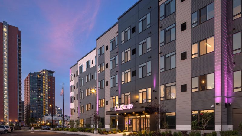 Westrum Corporate Modern apartment building with the sign "LUXOR MONTGOMERYVILLE" lit in purple, surrounded by city buildings at sunset.