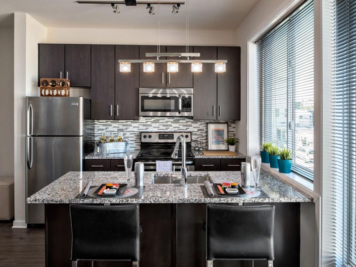 Westrum Corporate Modern kitchen at 1200 Washington with dark cabinets, granite island, stainless appliances, pendant lights, and a large window with blinds.