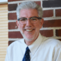Westrum Corporate Smiling man with gray hair and glasses, John J. Ernst, AIA, CBO, wearing a white shirt and blue tie stands before a brick wall.