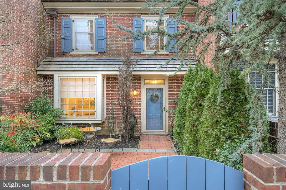 Westrum Corporate Willowbrooke brick townhouse with blue door, wreath, blue shutters, garden seating, and lush greenery behind a low brick wall and blue gate.