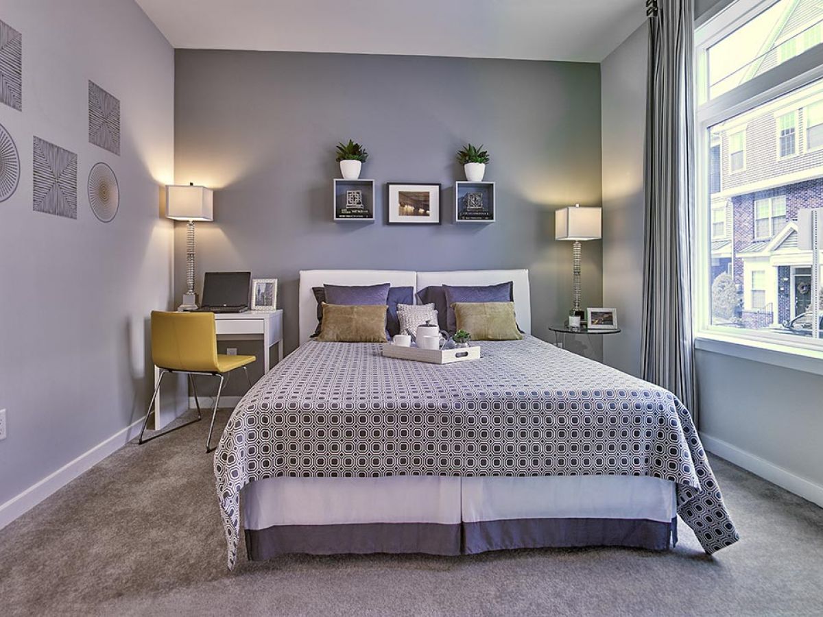Westrum Corporate Modern bedroom at Flats at 31 Brewerytown with patterned bedding, yellow chair, wall art, and large window overlooking Brewerytown apartments.