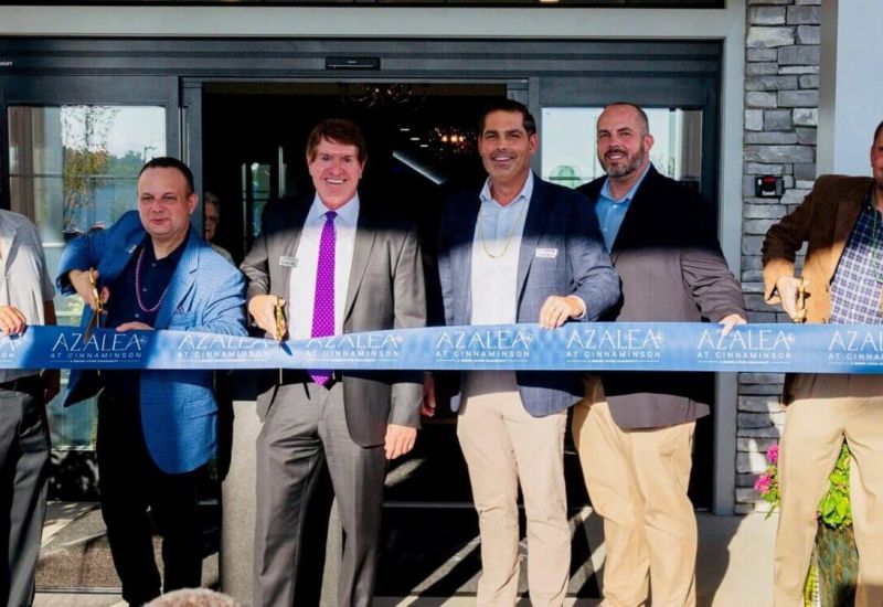 Westrum Corporate Eight people smile and hold a blue ribbon at a business ribbon-cutting ceremony in front of a building entrance.